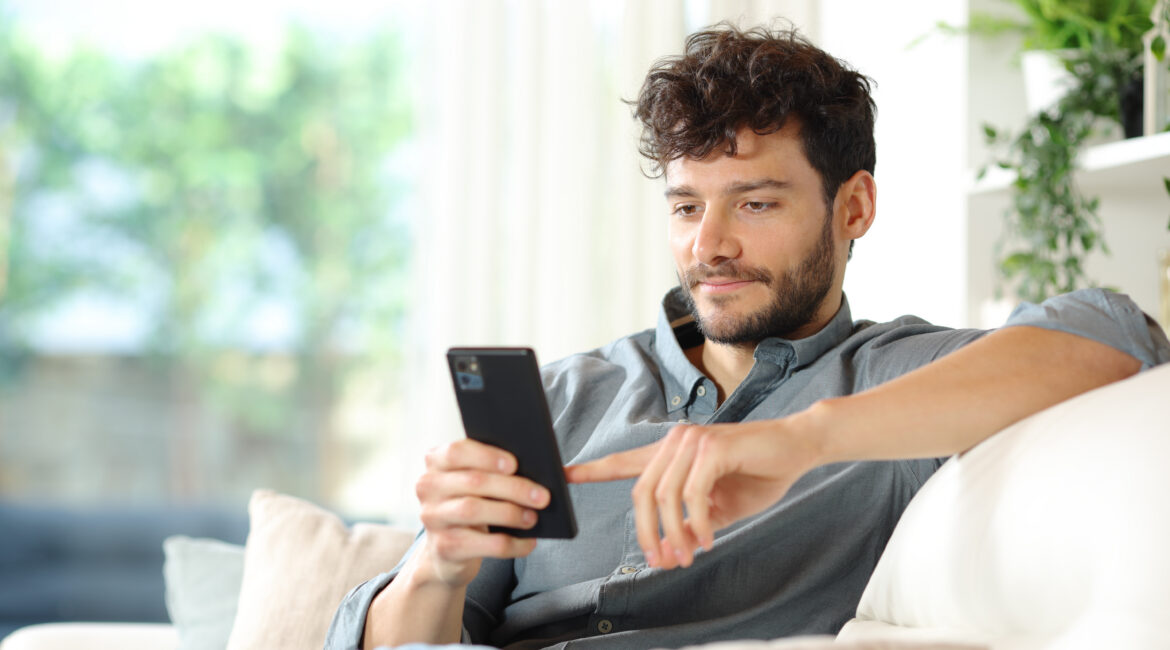 Man sitting on a couch at home in natural light, looking at a phone.