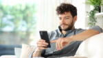 Man sitting on a couch at home in natural light, looking at a phone.