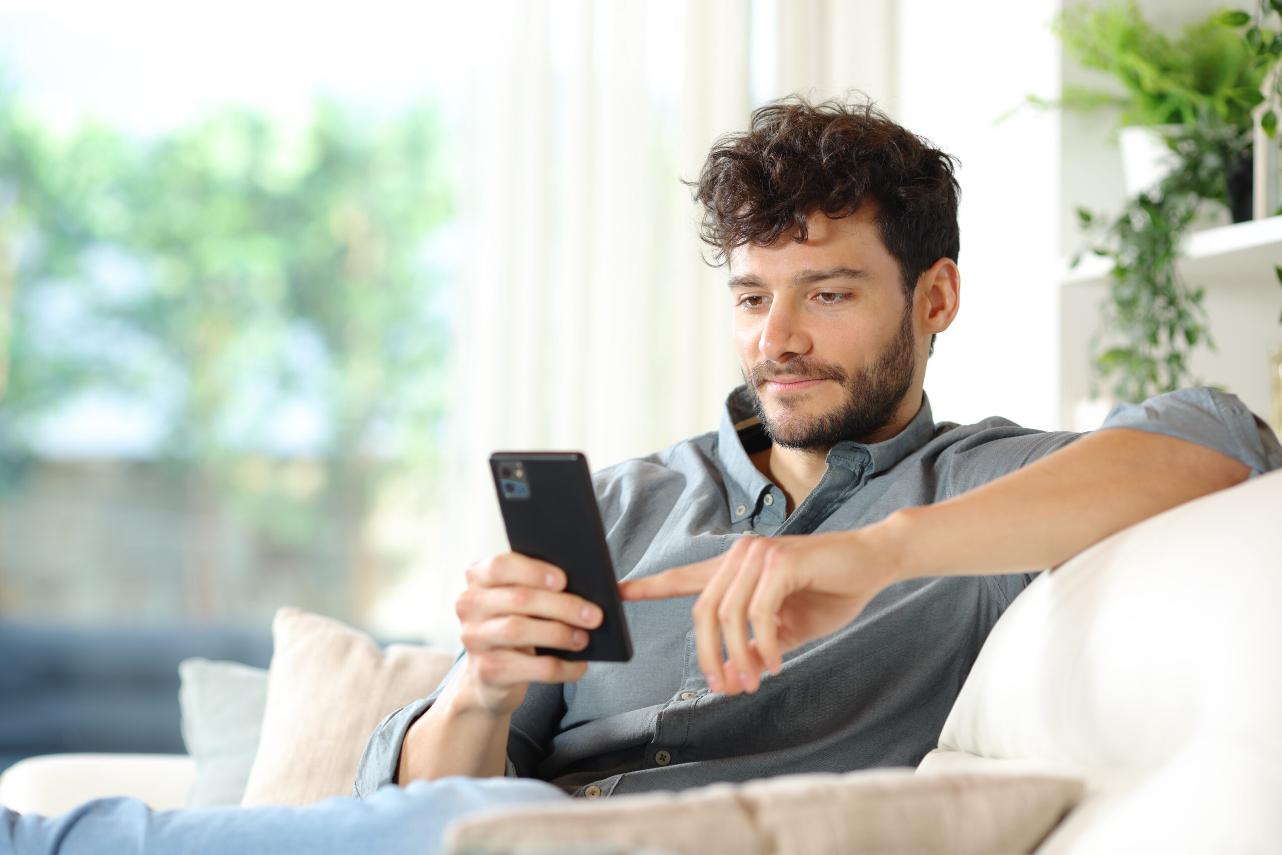 Man sitting on a couch at home in natural light, looking at a phone.